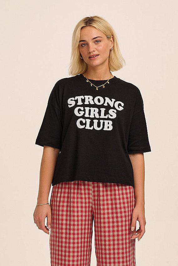 Woman wearing a black 'STRONG GIRLS CLUB' t-shirt and red checkered pants on a beige background