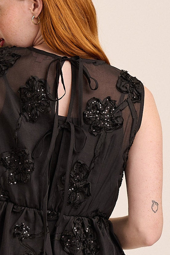 A woman with red hair is shown from the back wearing a sleeveless black tiered peplum top with floral lace applique and black pants, set against a plain beige studio background.