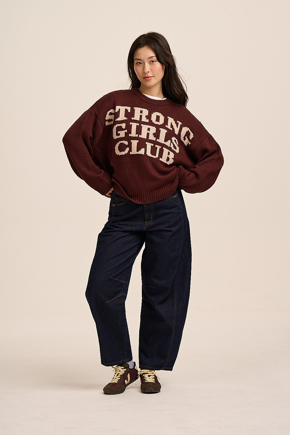 A woman modeling a dark maroon knit sweater with 'STRONG GIRLS CLUB' printed in cream across the chest, paired with dark blue jeans and brown sneakers, against a solid light beige background.