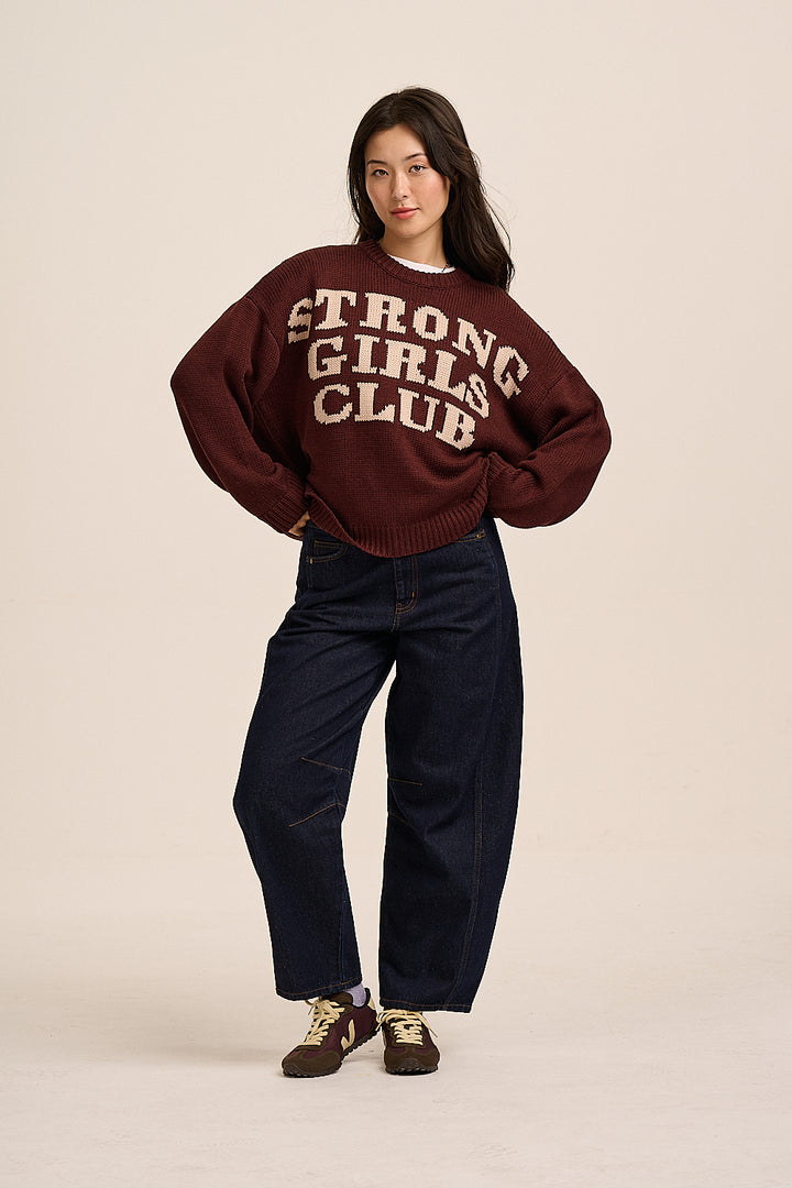 A woman modeling a dark maroon knit sweater with 'STRONG GIRLS CLUB' printed in cream across the chest, paired with dark blue jeans and brown sneakers, against a solid light beige background.