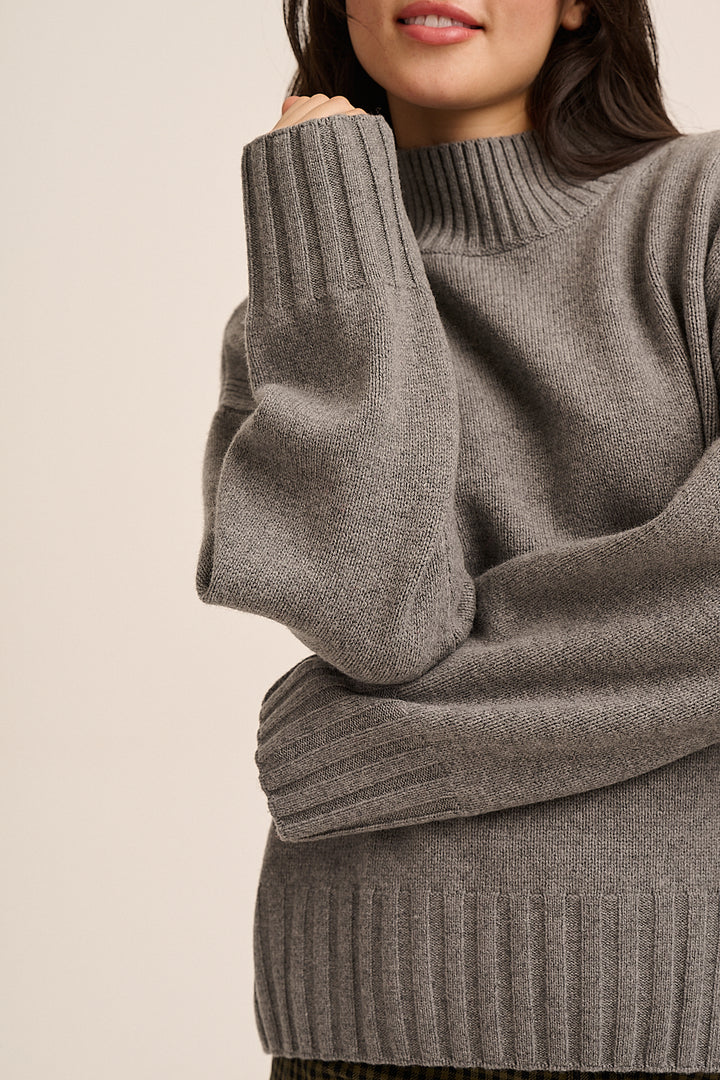 A close-up of a woman wearing a chunky ribbed knit sweater in a heather gray color, showing the texture and fit against a plain beige background.
