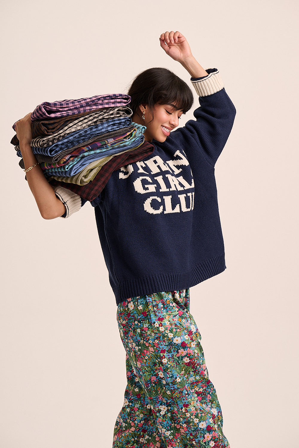 A woman modelling a navy blue knit sweater with 'STRONG GIRLS CLUB' in white varsity lettering, paired with a colourful floral skirt and carrying a stack of folded patterned blankets over her shoulder, against a solid light beige background.