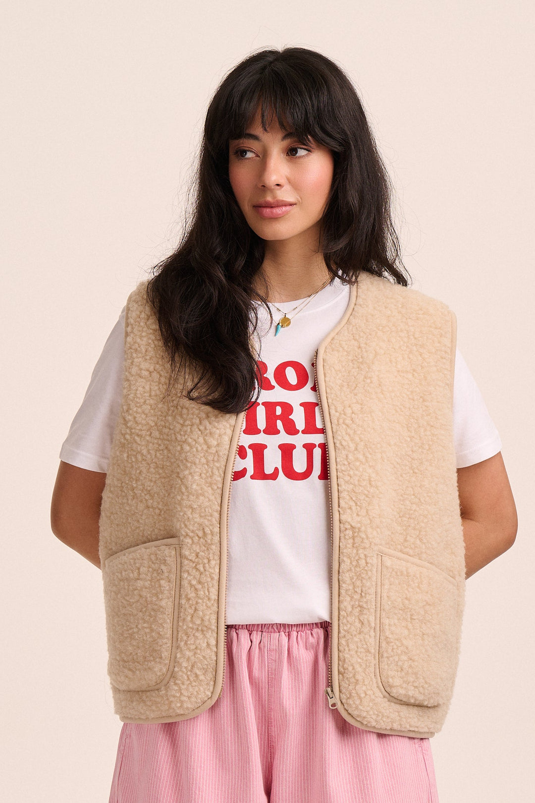 Person wearing a beige vest, white t-shirt with red text, and pink pants on a beige background